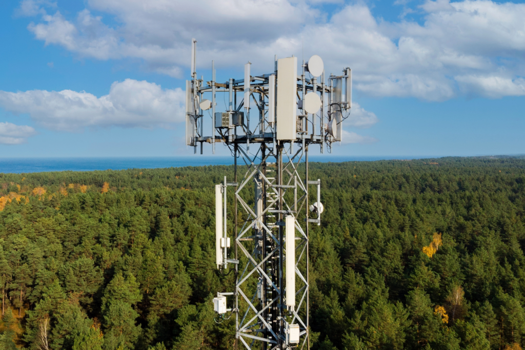 Telecommunications tower with panel antennas and microwave dishes above a wooded landscape.